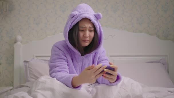 Αναστατωμένη Ασιάτισσα που παίζει βιντεοπαιχνίδια στο κινητό και χάνει το παιχνίδι καθισμένη στο κρεβάτι. Η κυρία έχασε το παιχνίδι στο smartphone. — Αρχείο Βίντεο