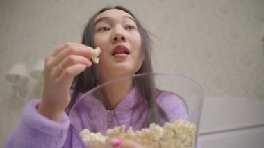 Asyalı bir kadının komik film ya da televizyon izlerken patlamış mısır yediği yakın plan. Mor pijamalı bir kadın televizyonun önünde gülüyor..