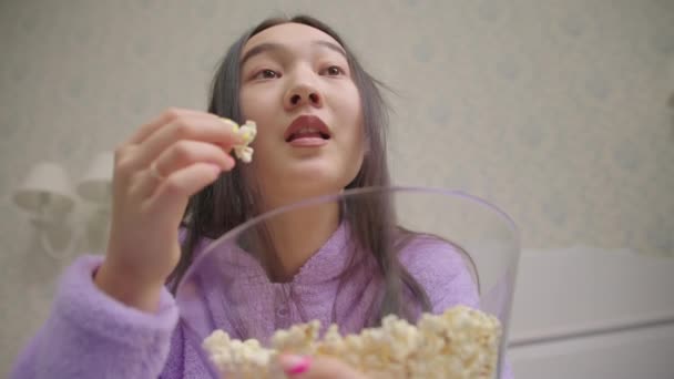 Close-up van Aziatische vrouw eten popcorn tijdens het kijken naar grappige film of tv-show. dame in paarse pyjama lachen in de voorkant van tv. — Stockvideo