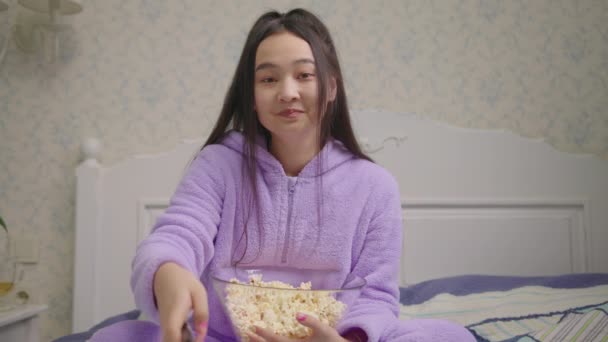 Mujer asiática viendo la película divertida o programa de televisión y comer palomitas de maíz sentado solo en la cama. Mujer en pijama púrpura riendo viendo comedia mirando a la cámara. — Vídeo de stock