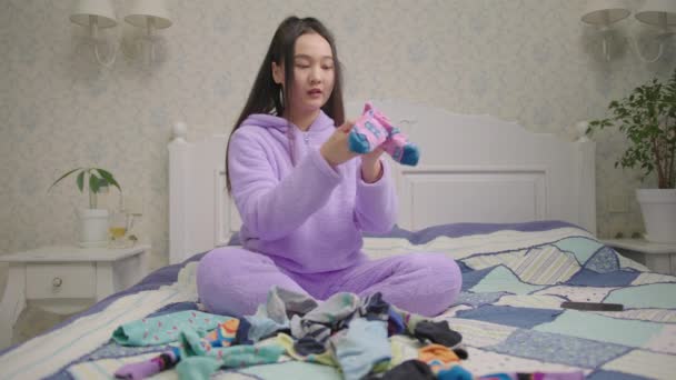 Jeune femme asiatique adulte pliant chaussettes de couleur assis sur le lit. Femme en pyjama violet organisant et simplifiant la maison en faisant l'ordre dans les vêtements. Vous cherchez une paire de chaussettes. — Video