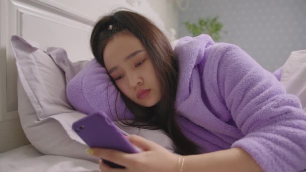 20s Asian woman browsing online using mobile phone lying in bed in the morning. Close up of woman in purple pajamas surfing web using cell phone in bed. — Stock Video