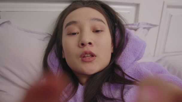ベッドに寝そべっているアジア人女性の笑顔の電話カメラハメ撮り。携帯電話を使って紫のパジャマを着た女性. — ストック動画