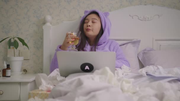 Kranke Asiatin der 20er Jahre arbeitet von zu Hause aus, trinkt heißen Tee und hustet im Bett liegend. Berufstätige Frau im lila Pyjama ist krank. — Stockvideo