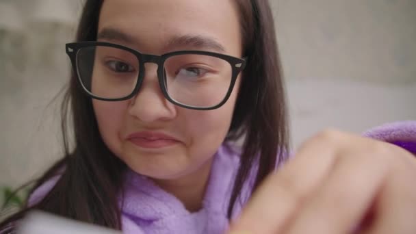 Gros plan de sortie asiatique femme en lunettes de lecture livre intéressant assis dans le lit. Caméra de poche, ralenti. — Video