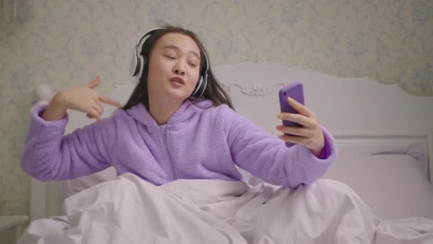 Азійська жінка в бездротовому навушнику співає за допомогою мобільного телефону. Щаслива жінка в пурпуровій піжамі співає і танцює з руками на ліжку.. — стокове відео