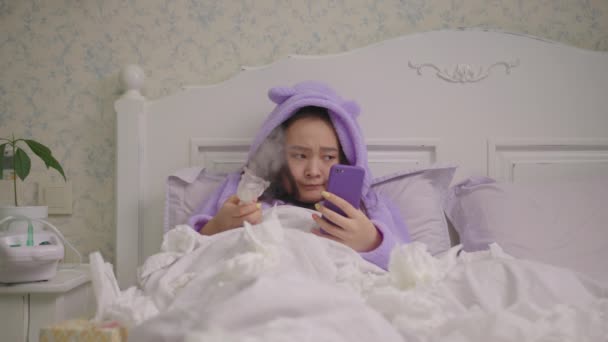 Millennial zieke Aziatische vrouw met behulp van mobiele telefoon in plaats van het gebruik van inhalator voor griep therapie in bed. Vrouwelijke behandeling van hoest met medicijninhalator en surfnet thuis. — Stockvideo
