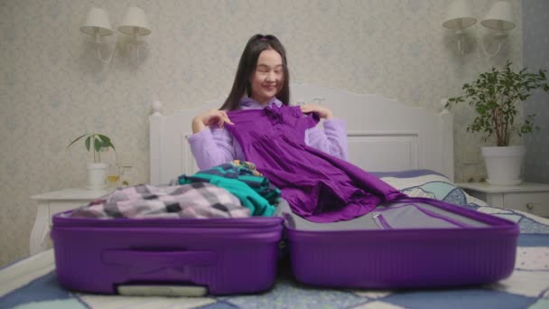 Šťastná asijská žena balení kufr na dovolenou sedí v ložnici sám. 20s žena sní o nadcházející dovolené držení barevných šatů. — Stock video