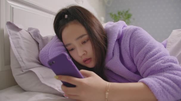 20s Asian woman browsing online using mobile phone lying in bed in the morning. Close up of woman in purple pajamas surfing web using cell phone in bed. — Stock Video