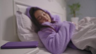 20 'li yaşlarda mor pijamalı Asyalı bir kadın uyanıyor ve elinde cep telefonuyla yatakta yatıyor. Uykulu kadın sabahları cep telefonu kullanıyor..