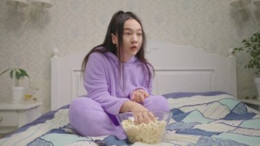 20 'li yaşlarda Asyalı bir kadın televizyon ya da film seyrediyor ve yatakta tek başına patlamış mısır yiyor. Mor pijamalı kadın TV karşısında korktu..