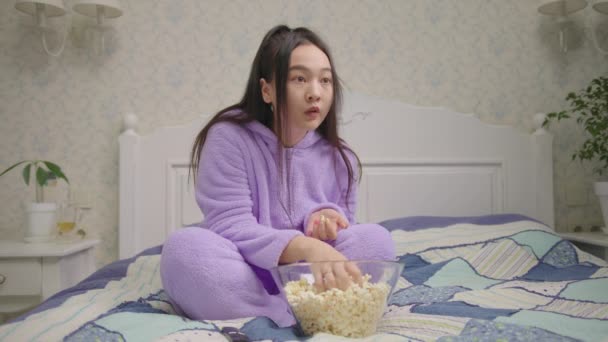 20s Asian woman watching exited movie or tv show and eating popcorn sitting alone on bed. Lady in purple pajamas got scared in front of tv. — Video Stock