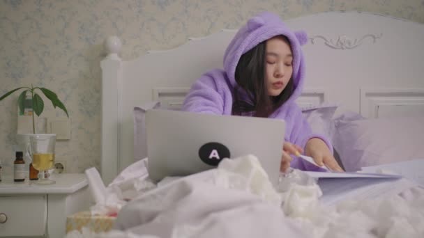 O femeie asiatică bolnavă de 20 de ani lucrează cu documente și laptop-uri întinse în pat. O femeie în pijamale violet senzație de frig. — Videoclip de stoc