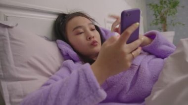 Yatakta cep telefonu kullanan dalgın Asyalı kadın. Cep telefonunda mesaj yazan kadın.