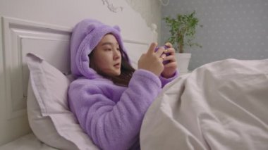 Genç Asyalı kadın cep telefonuyla video oyunu oynuyor ve yatakta yatarak oyunu kazanıyor. Bayan akıllı telefonda oynayarak kazanmaktan mutlu..