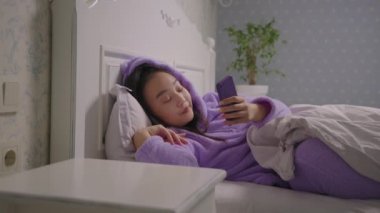 20 'li yaşlarda Asyalı bir kadın internette cep telefonuyla yatakta geziniyor. Mor pijamalı, gülümseyen kadın yatakta cep telefonu kullanarak internette sörf yapıyor..