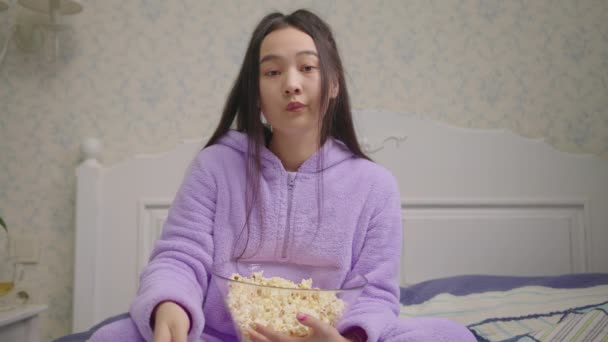 Znudzona Azjatka oglądająca telewizję i jedząca popcorn sama w łóżku. Kobieta z popcornem w domu patrząc na aparat pokazuje nudne emocje. — Wideo stockowe