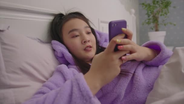 Feliz milenar mulher asiática digitando mensagem usando telefone celular deitado na cama. Mulher adulta jovem em pijama navegando on-line no telefone celular. Câmera portátil. — Vídeo de Stock