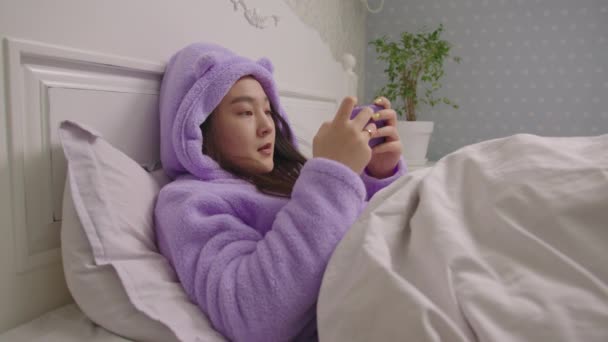Fiatal felnőtt ázsiai nő videojátékozik a mobiltelefonján, és megnyeri a játékot fekszik az ágyban. A hölgy szívesen nyer okostelefonon.. — Stock videók