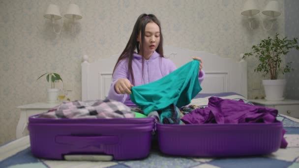 Millénaire asiatique femme emballage bagages assis sur le lit à la maison. Femme en pyjama à la recherche de vêtements en valise. — Video