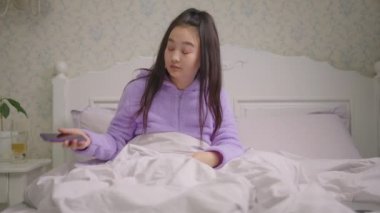 Uykulu 20 'li yaşlardaki Asyalı kadın internette cep telefonuyla geziniyor akşamları yatakta oturuyor. Mor pijamalı bir kadın yatakta cep telefonu kullanarak internette sörf yaparken esniyor..