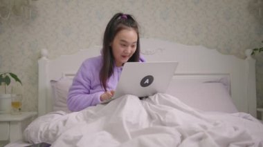 20 'li yaşlarda Asyalı bir kadın laptopta yatakta oturmuş iyi haberler okuyor. Milenyum kadınları mutlu ve dizüstü bilgisayar kullanarak çıkıyorlar. Mutlu kadın heyecan verici duygular gösterir..