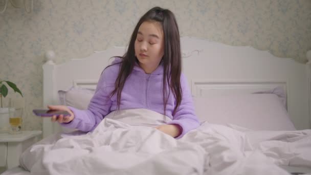 Slaperige jaren '20 Aziatische vrouw surfen op internet met behulp van mobiele telefoon zitten in bed in de avond. Vrouw in paarse pyjama gapen tijdens het surfen online met behulp van mobiele telefoon in bed. — Stockvideo