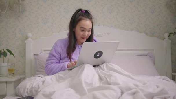 Dvacátnice Asiatka čte dobré zprávy na notebooku, jak sedí v posteli. Millennial žena je šťastná a opustil pomocí notebooku. Šťastná žena ukazuje wow emoce. — Stock video