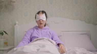 20 'li yaşlarda uyuyan uyku maskeli Asyalı bir kadın uyanıyor ve cep telefonuna bakıp geç kaldığını fark ediyor. Mor pijamalı kadın uyuyakalmış..