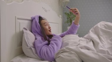 20 'li yaşlarda Asyalı bir kadın yatakta uzanmış cep telefonu kamerasına bakıyor. Mor pijamalı güzel kadın blogcu yataktan arıyor..