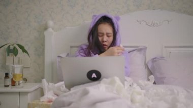 Sick Asian woman watching laptop lying in bed at home. Woman in purple pajamas spending time in bed with laptop watching comedy movies or tv show.
