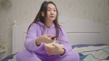 20 'li yaşlarda Asyalı bir kadın komik film ya da televizyon programı seyrediyor ve yatakta tek başına patlamış mısır yiyor. Mor pijamalı bir kadın televizyonun önünde gülüyor. Kapat..