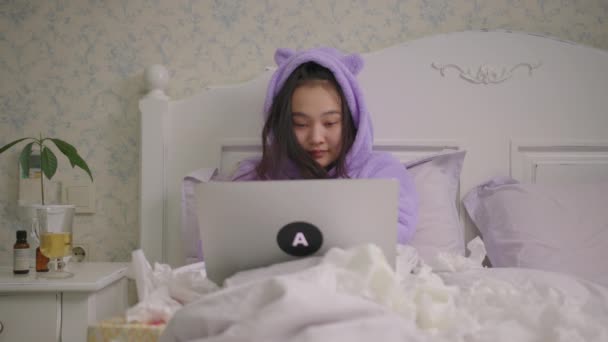 Sick Asian woman laughing watching laptop lying in bed at home. Woman in purple pajamas spending time in bed with laptop watching comedy movies or tv show. — стокове відео