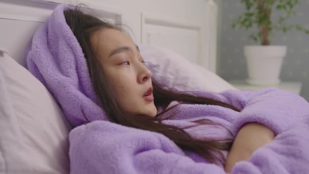 穿着紫色睡衣的疲惫的亚洲女人躺在床上，用羽绒被盖住自己。女人在家里睡着了. — 图库视频影像