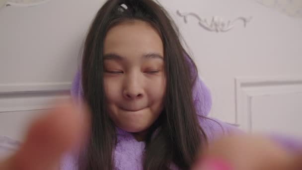 Cámara celular POV de mujer asiática sonriente escribiendo mensaje acostado en la cama. Mujer en pijama púrpura usando el teléfono celular desplazándose con el dedo en la pantalla. — Vídeos de Stock