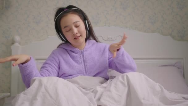 Milenyum Asyalı kadın, kablosuz kulaklıkla yatakta oturup müzik dinlemeyi sever. Mor pijamalı mutlu kadın elleriyle dans ediyor.. — Stok video