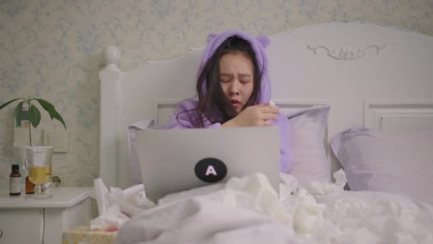 Sick Asian woman watching laptop lying in bed at home. Woman in purple pajamas spending time in bed with laptop watching comedy movies or tv show. — стоковое видео