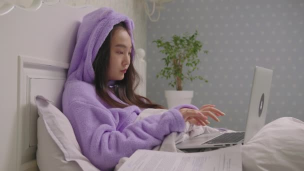 年轻的亚洲妇女在家里工作，她们使用笔记本电脑和躺在床上的文件。在家卧床的妇女远程工作. — 图库视频影像