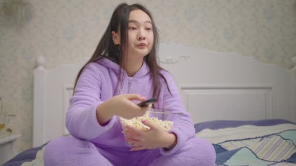 Aziatische vrouw uit de 20 die een grappige film of tv-show kijkt en popcorn eet die alleen op bed zit. Vrouw in paarse pyjama lachend voor de tv. Sluiten.. — Stockvideo