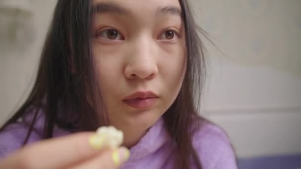 Primer plano de la mujer asiática comiendo palomitas de maíz mientras ve una película divertida o un programa de televisión. Dama en pijama púrpura riendo delante de la televisión. — Vídeo de stock