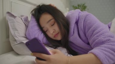 Uykulu 20 'li yaşlarda Asyalı bir kadın internette cep telefonuyla internette geziniyor ve akşamları yatakta yatıyor. Mor pijamalı bir kadının internette sörf yaparken yatakta akıllı telefon kullanarak esnediği yakın plan..