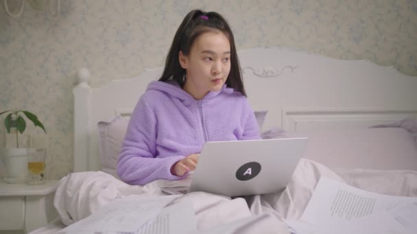 Millennial Asian woman exited working from home using laptop and documents sitting in bed. Female is happy to work remotely staying in bed at home. — Stock Video