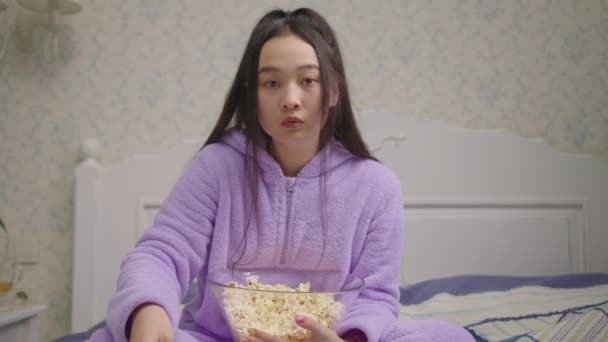 En asiatisk kvinna i 20-årsåldern som tittar på läskig film och äter popcorn ensam i sängen. Millennial kvinna med popcorn tittar på kameran och bli rädd. — Stockvideo