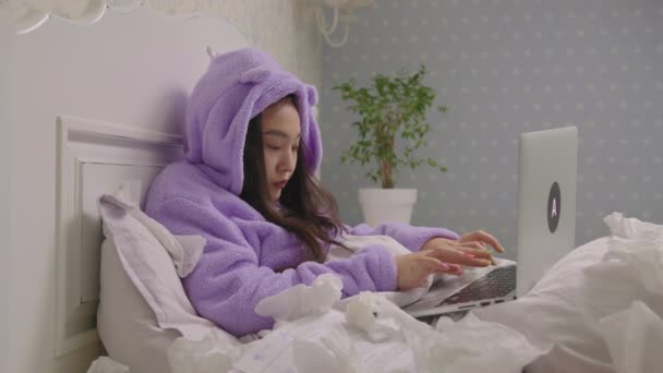 年轻的亚洲妇女打喷嚏咳嗽躺在床上在家工作。穿着紫色睡衣的生病女性在卧室的笔记本电脑上打字. — 图库视频影像