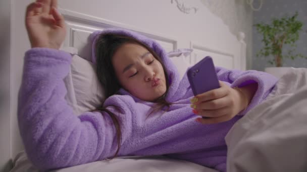 20s mujer asiática navegando en línea utilizando el teléfono móvil acostado en la cama. Primer plano de la mujer sonriente en pijama púrpura navegar por la web utilizando el teléfono celular en la cama. — Vídeos de Stock