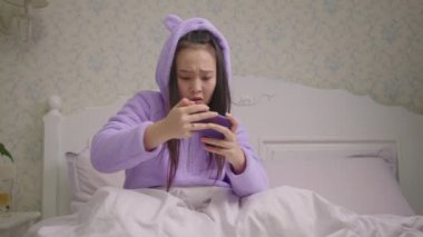 20 'li yaşlarda cep telefonuyla video oyunu oynayan Asyalı bir kadın yatakta oturup maçı kaybediyor. Kadın akıllı telefondan oyunu kaybetti..