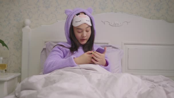 Sleepy 20s Asian woman in sleeping mask browsing internet using mobile phone sitting in bed in the evening. Woman in purple pajamas yawning while surfing online using cell phone in bed. — Stock Video