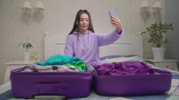 Glimlachende Aziatische vrouw pakt koffer en praat op mobiele telefoon videogesprek. Vrouw kijkt uit naar komende vakantie. — Stockvideo