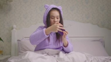 20 'li yaşlardaki Asyalı kadın cep telefonuyla video oyunu oynuyor ve oyunu yatakta oturarak kazanıyor. Akıllı telefonla oynarken zafer jesti yapan bir bayan. Kazandığıma sevindim..