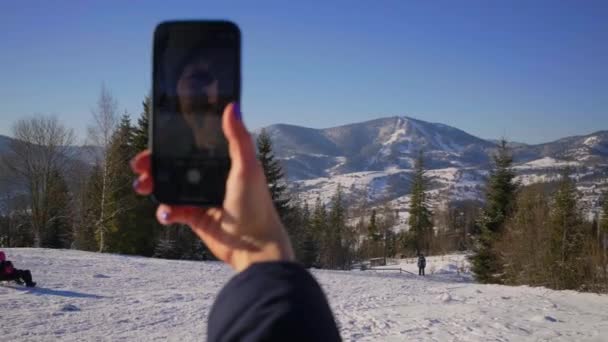 Kobieta ręka robi zdjęcie zimowego krajobrazu górskiego trzymając telefon komórkowy w rękach. Przez ramię widok osoby robiącej zdjęcie na komórce na zewnątrz. — Wideo stockowe